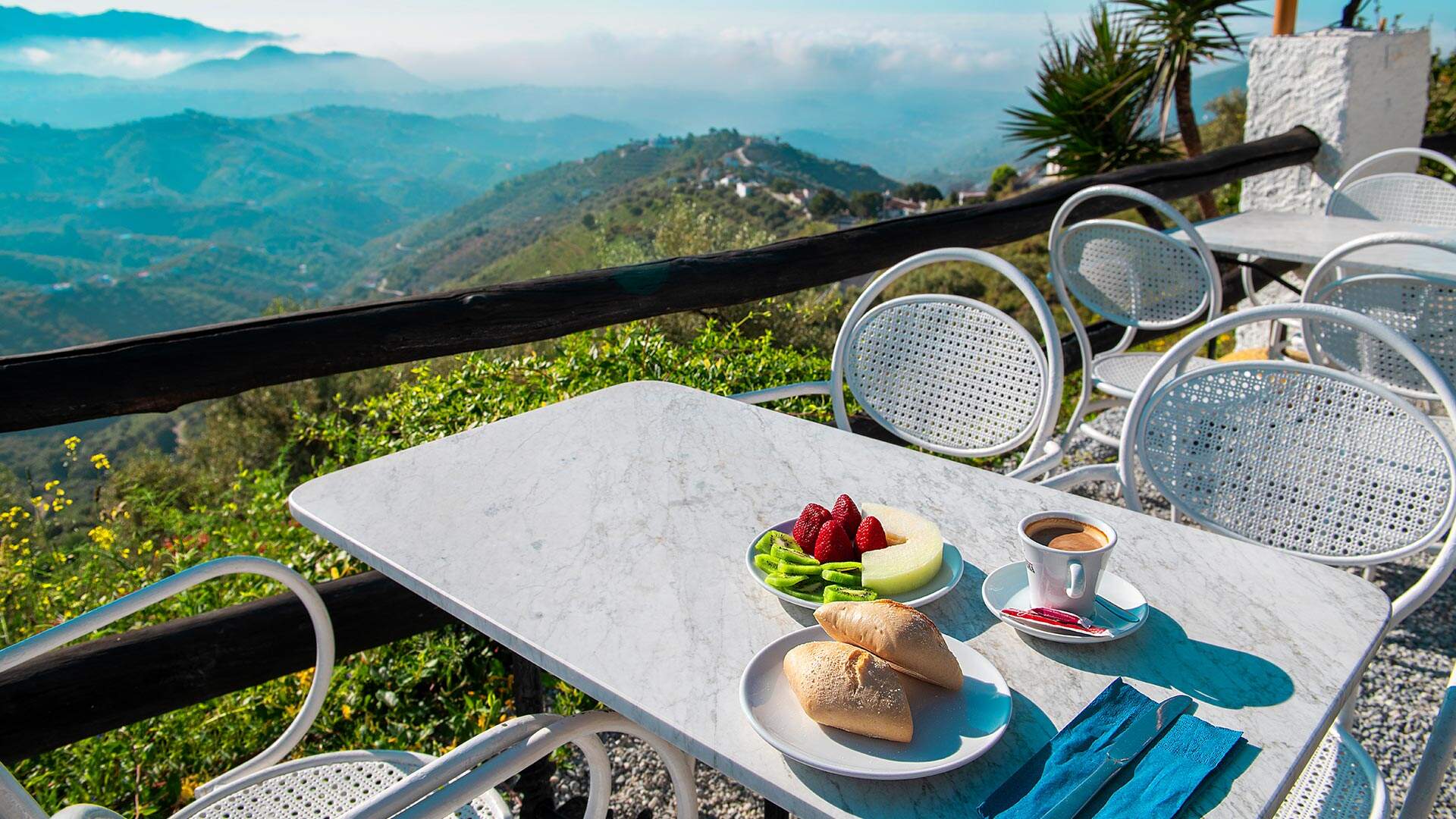 witte terrasstoeltjes met ontbijtje op tafel en uitzicht over hoge, mistige bergen