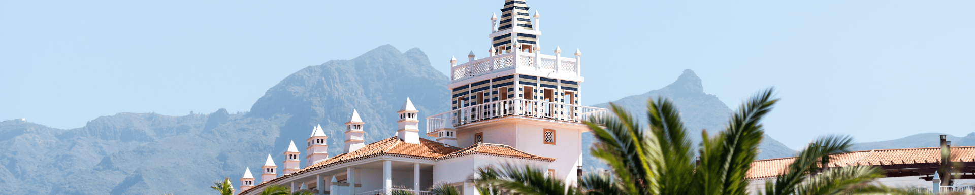 Uitzicht op een hotel op Tenerife met een kenmerkende toren en bergen op de achtergrond, omringd door palmbomen.